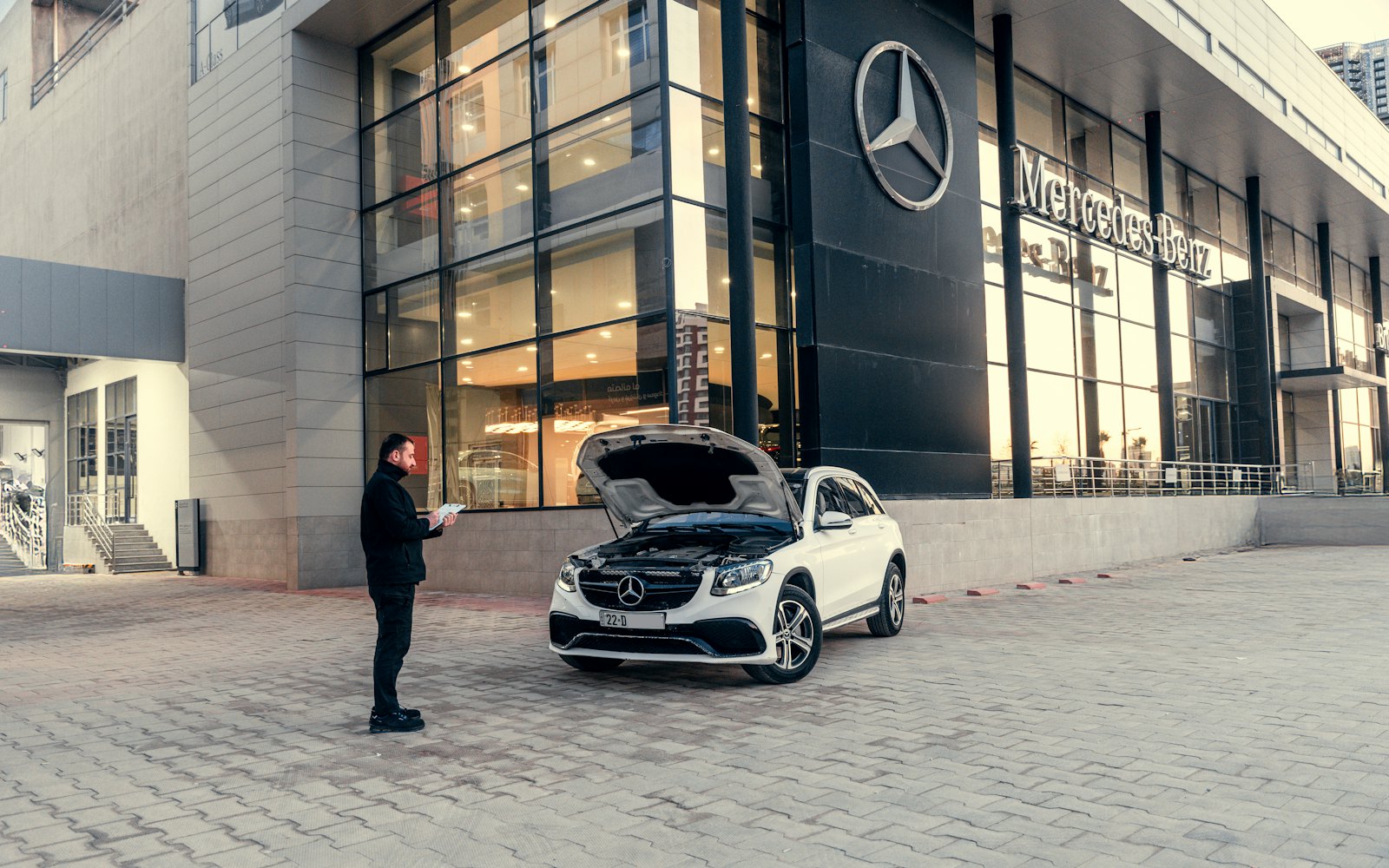 Salesperson with a clipboard inspecting a white SUV with its hood up outside a dealership showroom
