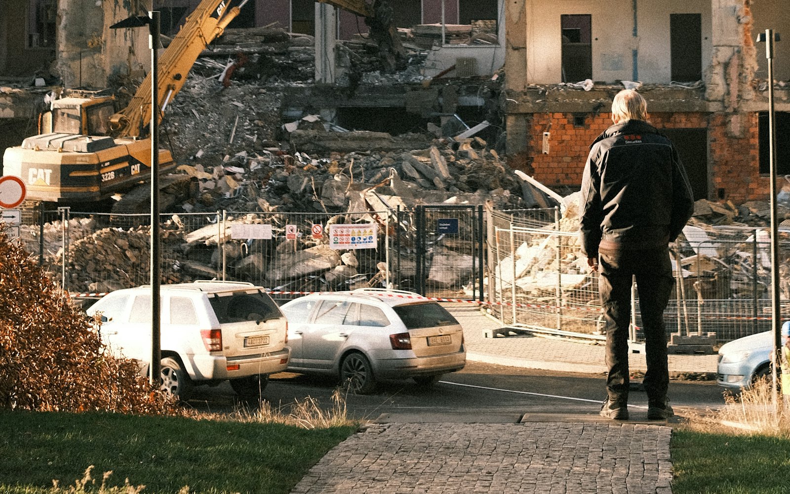 Excavator tearing down a concrete building behind site fencing while a worker watches from the street