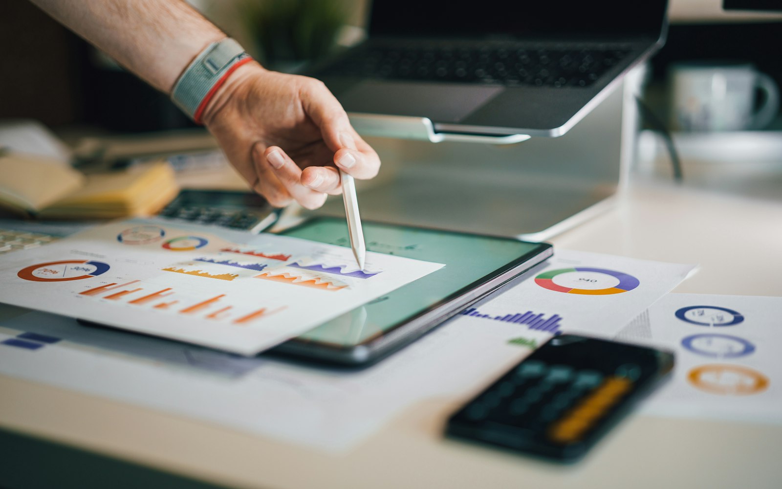 Hand with a stylus comparing marketing performance charts on a tablet and printed reports beside a laptop